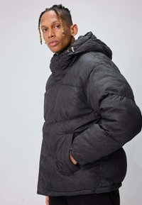 Veste matelassée noire avec un motif chevrons texturé, une capuche et des poches latérales. Le design est ample avec une fermeture éclair.