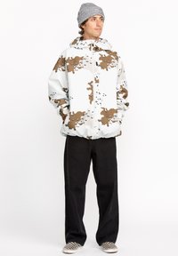 Witte jas met bruin abstract patroon, ritsluiting, en trekkoord capuchon; gecombineerd met zwarte broek en geblokte instappers.