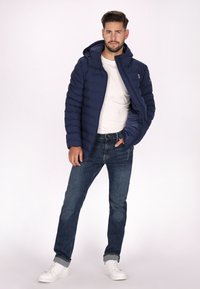 Mörkblå puffjacka med huva, tillverkad av vadderat material, kombinerad med en vit T-shirt och mörka jeans, kompletterad med vita sneakers.