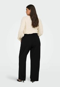 Frau mit langen dunklen Haaren, die sich abwendet. Sie trägt einen cremefarbenen Pullover mit Zopfmuster, schwarze weit geschnittene Hosen und schwarze hohe Schuhe.
