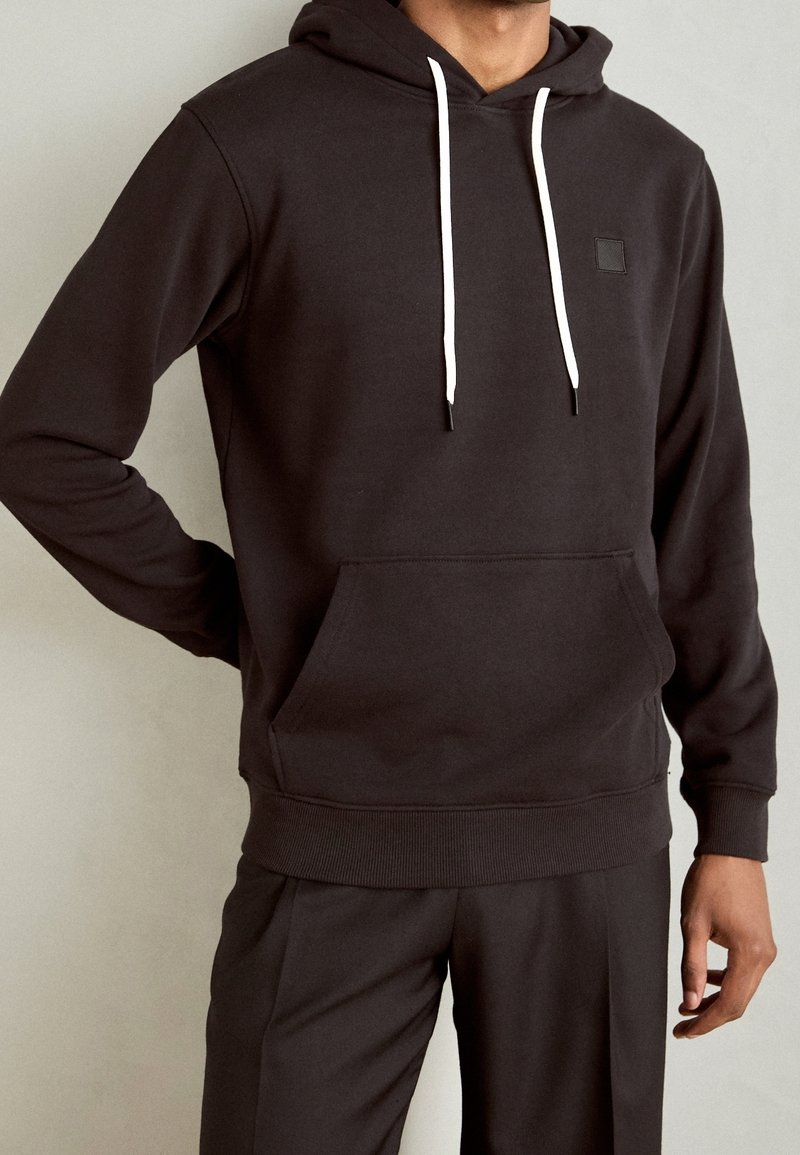 Sudadera de algodón gris oscuro con un bolsillo frontal, cordones blancos y una pequeña etiqueta negra en el pecho. Combinada con pantalones a juego.