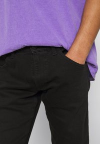 Svarta bomullsbukser med framfickor, tillsammans med en löst sittande lila t-shirt. Tyget verkar slätt med en lätt glans.