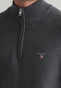 Sweatshirt zippé gris foncé en tissu texturé, avec un col montant, le logo GANT, et un petit blason sur la poitrine.