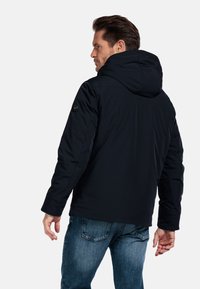 Marineblauwe hoodie-jack met een gladde textuur, voorzien van een zichtbaar logo op de mouw, gecombineerd met lichtblauwe spijkerbroek.