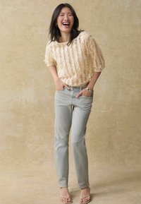 Haut court à manches courtes texturé beige clair, associé à un jean droit bleu clair et des sandales à lanières beiges, sur un fond neutre.