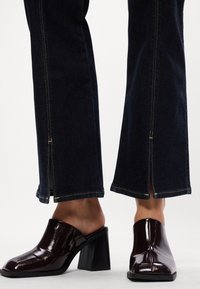 Rjavi patent usnjeni mule s kvadratnim prstom in blok peto, v kombinaciji z temno modrimi flare jeans hlačami, ki imajo stranske reže in kontrastne šive.