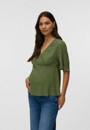 Gravid kvinde med langt mørkt hår iført en grøn tekstureret top og blå jeans, der blidt rører ved sin mave, stående foran en ensfarvet baggrund.