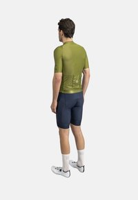 Maillot de cyclisme à manches courtes, de couleur vert olive avec une finition texturée, associé à un short bleu marine. Des chaussettes blanches et des chaussures de cyclisme complètent la tenue.
