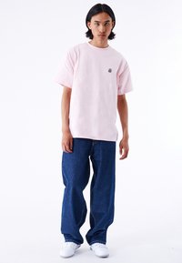 Camiseta de algodón rosa claro con mangas cortas y un pequeño logo negro, combinada con jeans de mezclilla azules holgados y zapatillas blancas.
