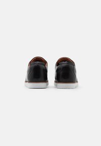 Scarpe in pelle nera con finitura liscia, dotate di suola in gomma bianca e contrasti marroni sul tallone. Solo vista posteriore.