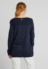 Marinblå långärmad topp med ett ljust mönster av små, färgglada motiv. Slät textur med en kontrasterande enfärgad panel vid axlarna.