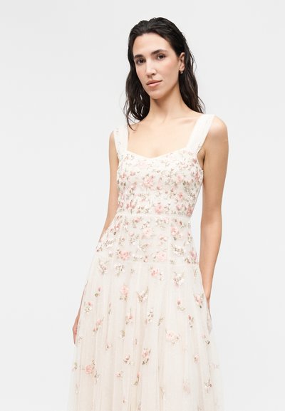 Γυναίκα με σκούρα μαλλιά φορά λευκό αμάνικο φόρεμα με ροζ και πράσινη κεντημένη λουλουδένια διακόσμηση σε απλό φωτεινό φόντο.