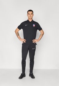 Sort Nike polo shirt med krave og Atlético Madrid logo, kombineret med sorte tapered bukser og sorte sportsko. Glat tekstur, tætsiddende design.