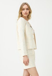 Tweed jasje en rokset in crème met een multicolore patroon. Jasje heeft een ronde hals en knoopsluiting, rok is aansluitend en komt tot boven de knie.