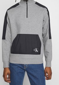 Felpa grigia con mezza zip, pannelli neri sulle spalle e tasca frontale. Presenta il logo di Calvin Klein Jeans sulla tasca.