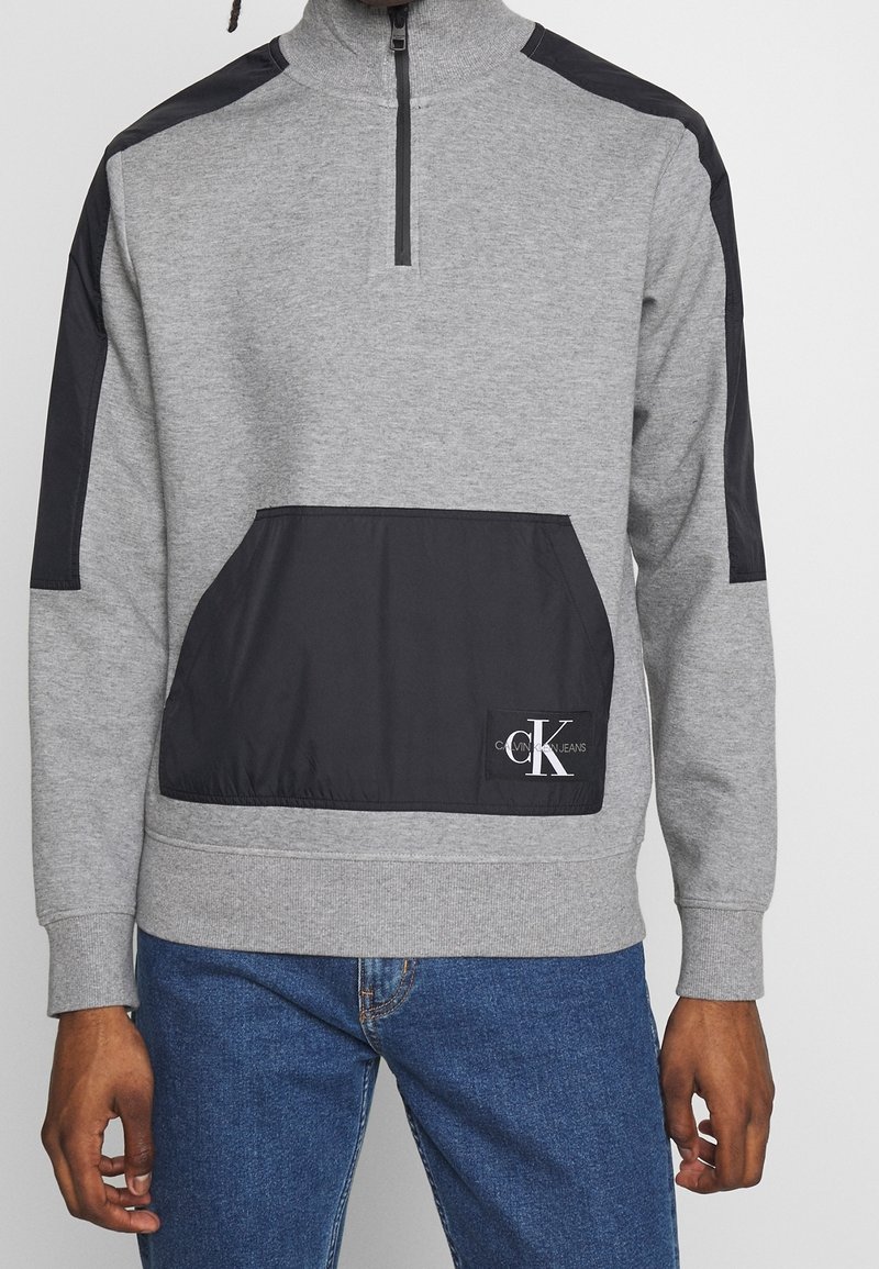 Felpa grigia con mezza zip, pannelli neri sulle spalle e tasca frontale. Presenta il logo di Calvin Klein Jeans sulla tasca.