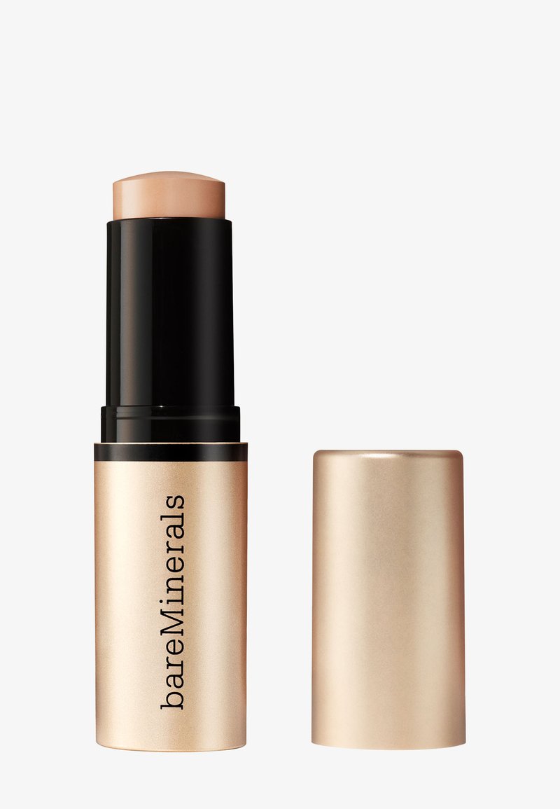 BareMinerals cream foundation stick with beige shade, black and gold casing, and matching gold cap placed beside it.