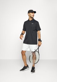 Schwarzes Polohemd mit Kragen und Nike-Logo, weiße Shorts, schwarze Sportschuhe und ein Tennisracket. Das Modell trägt eine schwarze Visor.