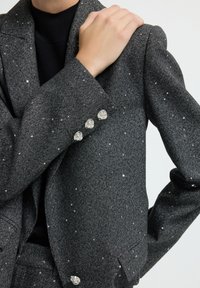Person in einem dunkelgrauen, melierten Blazer mit silbernen Knöpfen, die Hand ruht auf der gegenüberliegenden Schulter, schwarzes Oberteil ist darunter sichtbar.