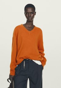 Orange Wolle Pullover mit V-Ausschnitt, lockerer Passform und gerippten Bündchen. Kombiniert mit navyblauen Nadelstreifenhosen und einer schwarzen Handtasche.