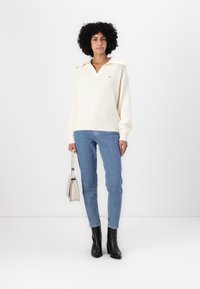 Krem farge pullover genser med kragehals, rillede teksturer, sammen med blå jeans, svarte hæle støvler og en liten hvit håndveske.