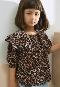 Blouse imprimée léopard avec col à volants et manches bouffantes, présentant des couleurs marron, noir et blanc ; des boutons en bois complètent le design.