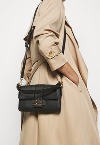 Mustapohjainen nahkalaukku, jossa on karkea pinta, kultaiset yksityiskohdat ja irrotettava hihna. Sisältää etutaskun ja logovarustuksen.