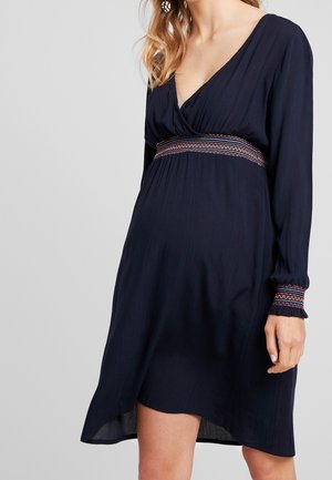 Kobieta ubrana w granatową sukienkę z długim rękawem z haftowanym pasem i mankietami, z dekoltem w serek i asymetrycznym obszyciem.