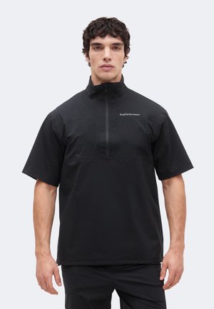 Mężczyzna w czarnym krótkim swetrze z półzamkiem i logo "Peak Performance" na lewej piersi na jednolitym tle.