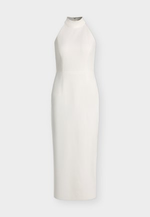 Vrouw in witte mouwloze midi-jurk, die een zwarte clutch vasthoudt, zwarte sandaaltjes met hak draagt, staand tegen een effen lichte achtergrond.