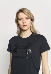 Juodas medvilnės marškinėliai su blizgia juoda logo ir tekstu "UNDER ARMOUR" krūtinėje. Apvalus kaklas ir trumpomis rankovėmis. Slydus tekstūra.