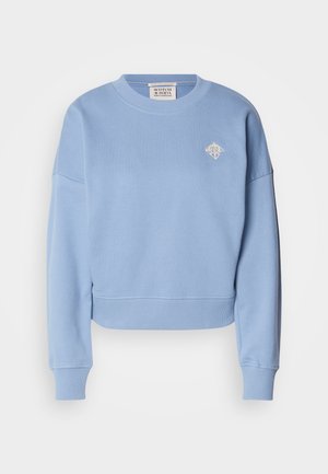 Lys blå kort genser laget av mykt materiale. Har rund hals, droppede skuldre og et lite brodert design på brystet.