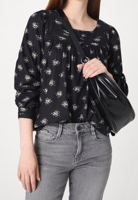 Femme portant un chemisier fleuri noir et un jean gris, tenant un sac à bandoulière en cuir noir contre sa poitrine.