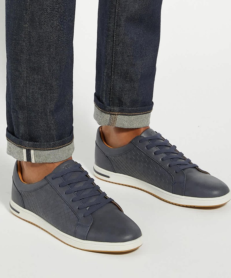 Pieds portant des baskets en cuir tressé bleu foncé et un jean en denim foncé retroussé sur fond blanc.