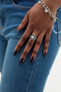 Anillo de plata con una piedra rosa en forma de corazón, diseño intrincado. Mano con uñas de un púrpura intenso reposa sobre unos jeans de mezclilla azul. Pulsera de cadena visible.