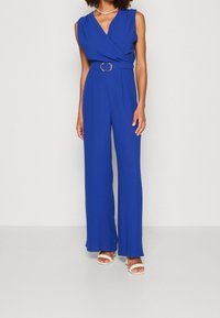 Blå ärmlös byxdress med en omslagdesign, vida byxor, midja med bälte och rektangulär spänne, samt en texturerad tygfinish.