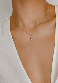 Collana d'oro con pendente a forma di rosa, caratterizzata da dettagli intricati e una catena sottile, indossata su una clavicola nuda e tessuto bianco.