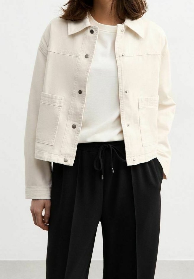 Femme portant une veste blanche boutonnée sur une chemise blanche avec un pantalon noir à cordon, debout devant un fond uni.