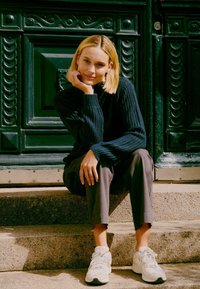 Sweter w kolorze granatowym, szare spodnie w wersji cropped i jasne sneakersy. Siedzi na schodach z zielonym, ozdobnym drzwiami w tle. Miękkie światło podkreśla fakturę.