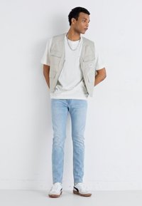 Light gray vest with multiple pockets over a white t-shirt, paired with light blue jeans and white sneakers with dark accents.