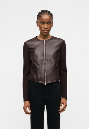 Woman with braided hair wearing a fitted dark brown leather jacket with front zipper and black pants, standing against a plain background.