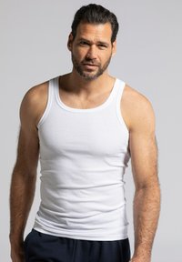 Weißes, geripptes Tanktop aus Baumwolle mit rundem Ausschnitt und breiten Trägern. Kombiniert mit dunklen Jogginghosen. Das Model hat kurze Haare und einen Bart.