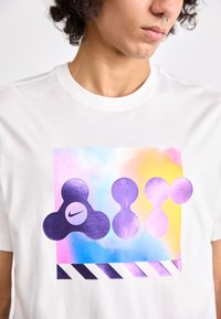 Λευκό T-shirt με πολύχρωμο γραφικό που απεικονίζει αφηρημένα σχήματα σε μωβ, ένα λογότυπο Nike και μια ριγέ λεπτομέρεια στο κάτω μέρος.