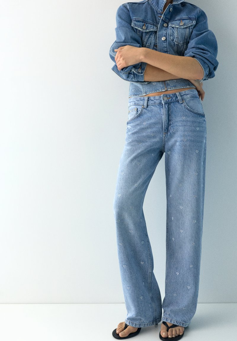 Personne portant un jean large brodé bleu clair, une veste en jean avec les manches retroussées, debout les bras croisés et pieds nus dans des sandales noires.