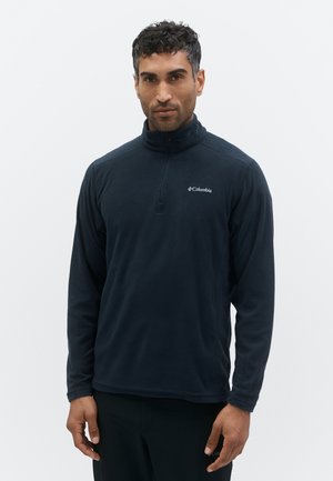 Vīrietis, tērpies melnā Columbia vilnas džemperī ar ceturtdaļas rāvējslēdzēju, stāv pret vienkāršu gaišu fonu.