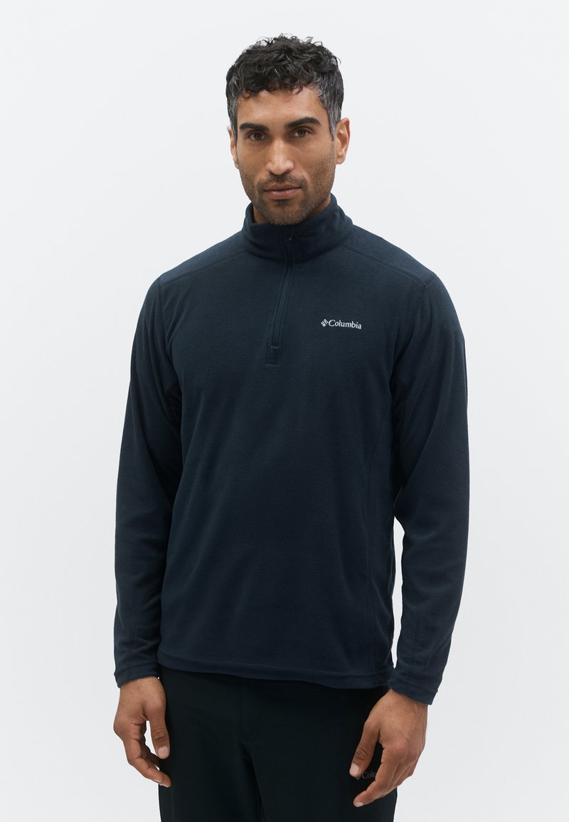 Man draagt een zwarte Columbia fleece met een kwartrits en staat tegen een effen lichte achtergrond.