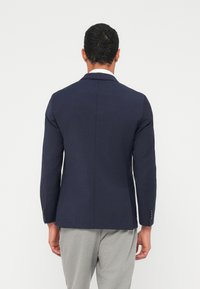 Ναυτικό μπλέ σακάκι με εφαρμοστή γραμμή, γιακάδες με σχισμές και δύο κουμπιά, κατασκευασμένο από λείο, ελαφρύ ύφασμα, συνδυασμένο με γκρι παντελόνια.