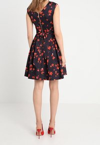 Frau in einem ärmellosen marineblauen Kleid mit rotem Blumenmuster, Reißverschluss auf der Rückseite und gebundenem Taillengürtel, kombiniert mit roten High Heels.