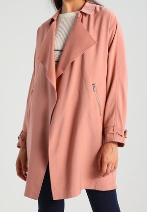 Frau trägt einen lockeren, staubrosa Trenchcoat mit breiten Revers, Reißverschlusstaschen an den Seiten und Schnallenriemen an den Ärmeln über einem leichten Pullover.