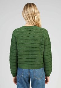 Pull en tricot vert avec un motif de mailles ouvertes texturées, à encolure ronde et poignets côtelés, associé à un jean en denim bleu.